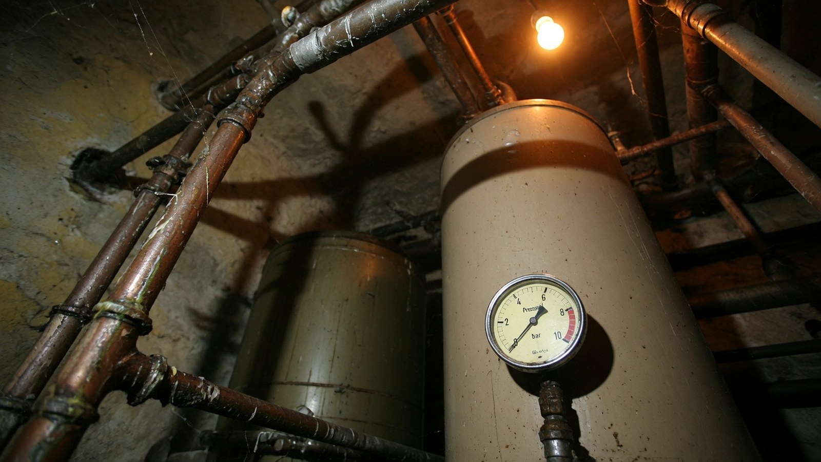 Ancienne chaudière au mazout beige en cave de pierre, tuyauterie rouillée au premier plan, réservoir de fuel flou en arrière-