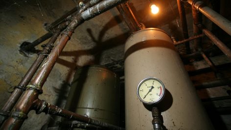 Ancienne chaudière au mazout beige en cave de pierre, tuyauterie rouillée au premier plan, réservoir de fuel flou en arrière-